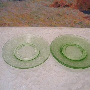 May include: Two green glass plates with a decorative floral pattern. The plates are round with a slightly raised center and a delicate, etched design. The plates are on a white lace tablecloth.
