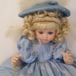 May include: A porcelain doll wearing a blue dress and hat with white feathers. The doll has blonde curly hair and blue eyes. She is holding a gold necklace with a heart pendant.