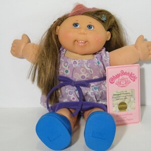 May include: A Cabbage Patch Kids doll with long brown hair, blue eyes, and rosy cheeks. The doll wears a purple dress with a matching bow and blue shoes. A pink birth certificate is included.