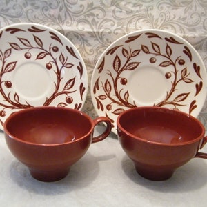 May include: Two brown teacups with white saucers. The saucers have a brown floral design with leaves and berries.