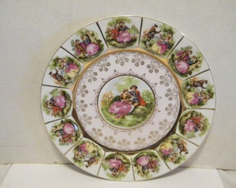 May include: A white porcelain plate with a gold rim and a central design of a couple in a garden. The plate is decorated with 12 smaller scenes of couples in various settings, all in a romantic style.