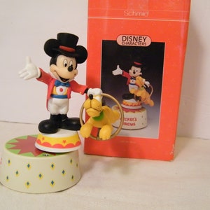 May include: A musical figurine of Mickey Mouse dressed as a ringmaster with Pluto jumping through a hoop. The figurine is on a circular base with a red, yellow, and white circus tent design. The box is red with the words "Disney Characters" and "Mickey's Circus" printed on it.