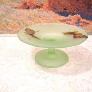 May include: A light green, frosted glass pedestal dish. The dish has a round base and a shallow, circular bowl. Two dragonfly designs are painted on the bowl in brown and yellow. The dish is likely vintage and decorative.