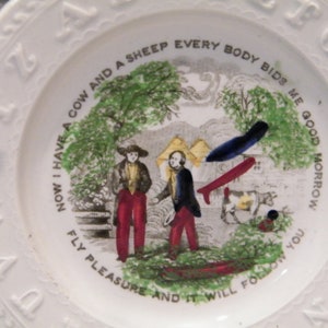 Peut inclure: Une assiette en céramique blanche avec une bordure bleue et rouge et une illustration verte, jaune et marron de deux personnes, une vache et un mouton. L'assiette porte le texte "Now I have a cow and a sheep every body bids me good morrow fly pleasure and it will follow you" autour du bord.