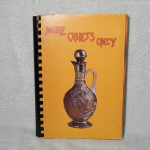 May include: A cookbook titled "More Crusts Only" with a spiral binding. The cover is orange and features a decorative brown glass bottle with a silver stopper and handle. The bottle has a floral design.