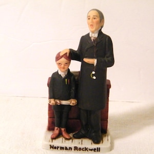 May include: A ceramic figurine depicting two figures, one adult and one child, in formal attire. The adult figure, in a long black coat, has one hand on the child's head. The child, wearing glasses, holds a scroll. The base reads "Norman Rockwell."