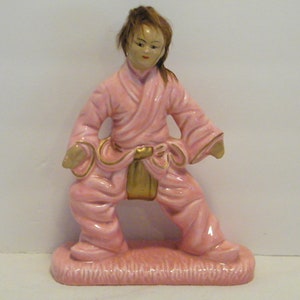 May include: A pink ceramic figurine of a person wearing a pink robe with a gold belt. The figurine is standing on a pink base with a textured surface.