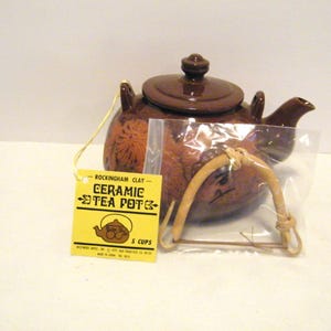 May include: A brown ceramic teapot with a lid, spout, and handle. The teapot has a floral design and a yellow tag that reads "Rockingham Clay Ceramic Tea Pot 5 Cups". The handle is in a plastic bag.