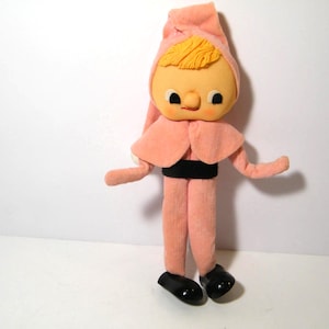 May include: A pink felt elf doll with a yellow yarn wig, black belt, and black shoes. The elf has a smiling face with black eyes and a large, round nose.
