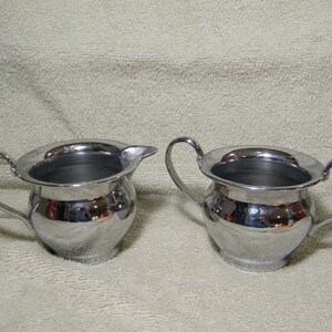 Peut inclure: Deux sucriers et crémier argentés brillants. Chaque récipient a un corps arrondi, une anse et une large ouverture. Le crémier a un bec verseur. L'ensemble est destiné à servir le café ou le thé.