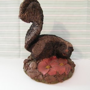 May include: A black and white skunk figurine with a curled tail, standing on a base with brown and red flowers. The base is decorated with small brown leaves and a snail.