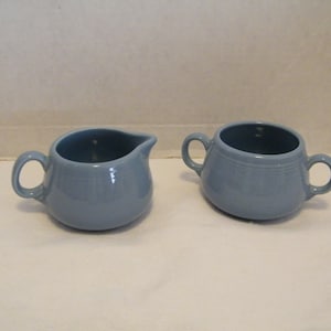 May include: A light blue ceramic creamer and sugar bowl set. The creamer has a spout and a handle. The sugar bowl has two handles. Both pieces have a rounded shape and are made of ceramic.