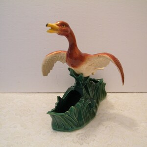 May include: A ceramic planter shaped like a duck with outstretched wings. The duck is brown and white with a yellow beak. The planter is green and has a leafy design.