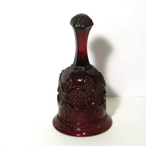 May include: A ruby red glass bell with a textured surface and intricate floral and geometric patterns. The bell has a long, slender neck and a detailed top. The bell is a decorative item.
