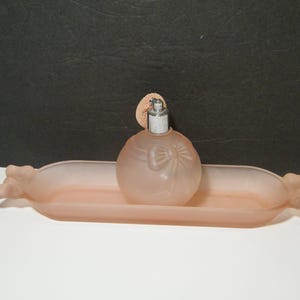 May include: A vintage, frosted pink perfume bottle with a bow design, topped with a silver atomizer and a pink mesh puff. It sits on a matching pink, oblong dish with decorative ends. The set is displayed against a dark background.