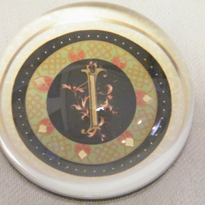 May include: A clear glass paperweight with a decorative design. The paperweight features a central black circle with a gold letter "I" and floral accents. The design is surrounded by green, red, and gold patterns, and a black border.