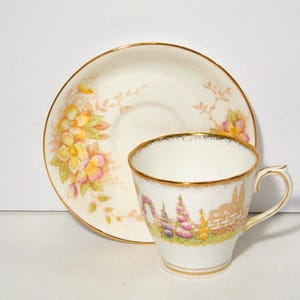 May include: A vintage teacup and saucer set. The cup and saucer are white with gold trim. The saucer features a floral design with yellow and pink flowers. The cup has a landscape scene with purple and pink flowers.