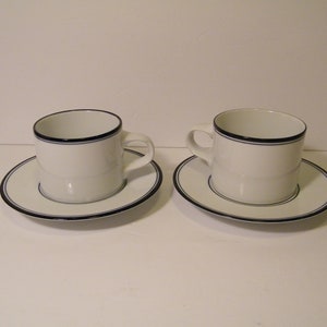 May include: Two white ceramic coffee cups with a black and blue trim. The cups are sitting on matching saucers.