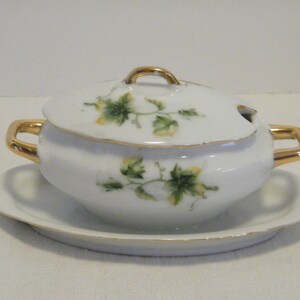 May include: White porcelain gravy boat with a gold trim and a lid. The boat has a floral design with green leaves and yellow flowers. It comes with a matching saucer.
