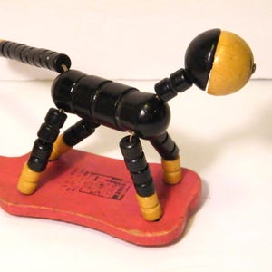 May include: A vintage wooden dog toy with a black body and yellow accents. The dog is articulated and mounted on a red wooden base. The head is half black and half yellow. The toy is a classic example of folk art.