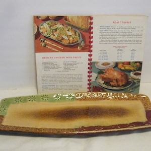 May include: A vintage cookbook with a red and white cover, open to a recipe for Mexican Chicken with Fruits. The cookbook is lying on a rectangular ceramic serving dish with a brown, green, and white glaze.
