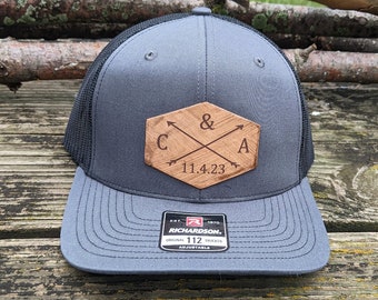 May include: A gray and black trucker hat with a wooden patch that says "C & A 11.4.23" with crossed arrows. The hat is adjustable and has a Richardson 112 label.
