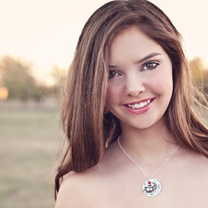 May include: A silver necklace with a circular pendant displaying a colourful illustration. The pendant hangs from a fine chain. The image shows a person with long brown hair, smiling, with a blurred outdoor backdrop.