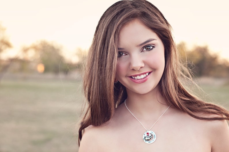 May include: A silver necklace with a circular pendant displaying a colourful illustration. The pendant hangs from a fine chain. The image shows a person with long brown hair, smiling, with a blurred outdoor backdrop.