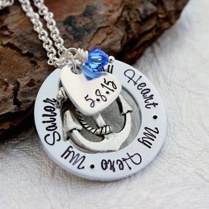 May include: A silver necklace with a pendant featuring an anchor, heart, and the words "Sailor My Heart My Hero." A blue crystal bead accents the top. The date "5.8.15" is inscribed on the heart. The necklace is on a silver chain.