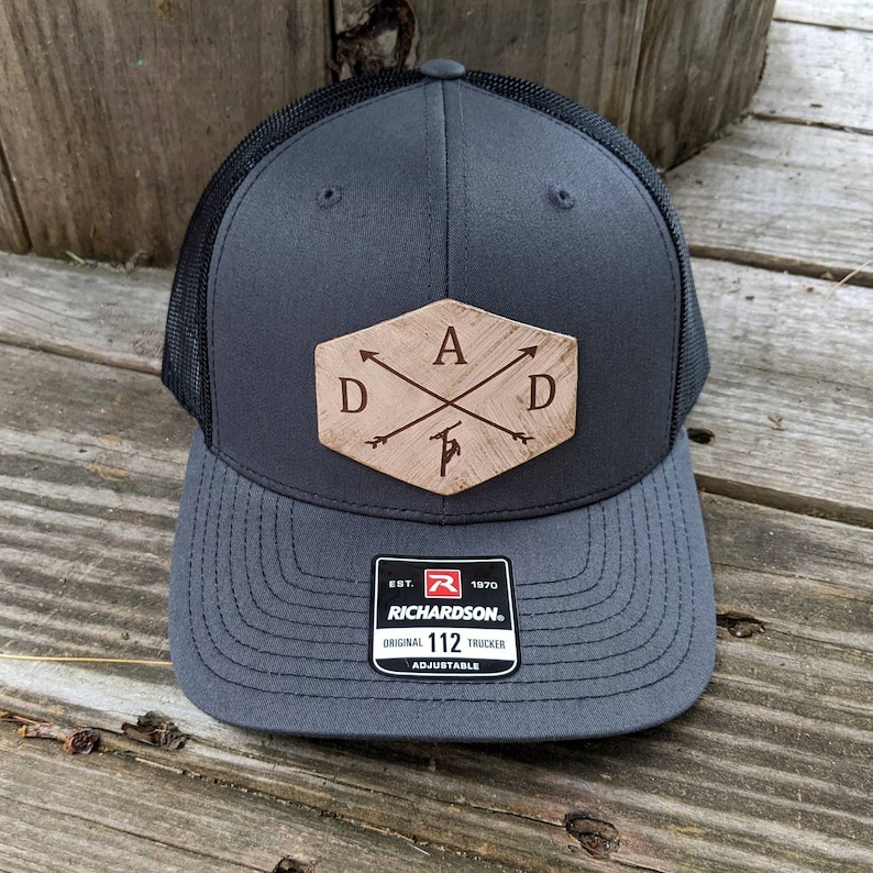 K&ouml;nnte beinhalten: Eine graue und schwarze Trucker-Kappe mit einem braunen Leder-Patch, auf dem "DAD" mit Pfeilen steht, die auf die Buchstaben zeigen. Der Patch hat die Form eines Sechsecks. Die Kappe hat ein schwarzes Netzr&uuml;cken und eine graue Vorderseite. Die Kappe ist verstellbar und hat ein Richardson 112-Etikett.
