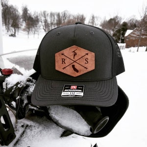 May include: Black trucker hat with a brown leather patch featuring a stylized logo with the letters "R" and "S" and a silhouette of a person holding a tool. The patch also includes a small outline of the state of California. The hat has a black mesh back and a black brim with the text "EST. 1970 RICHARDSON. ORIGINAL 112 TRUCKER. ONE SIZE" printed on a white label.