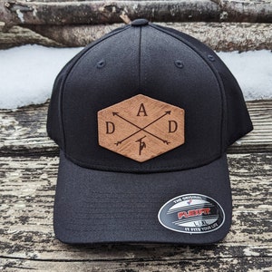 Peut inclure: Casquette de baseball noire avec un patch en cuir marron représentant les lettres "DAD" avec un motif de flèche stylisé et la lettre "A" au centre. Le patch a une forme hexagonale. La casquette a une étiquette Flexfit au dos.