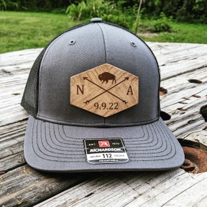 May include: A gray and black trucker hat with a wooden patch that says "N.A. 9.9.22" with a buffalo silhouette and crossed arrows. The hat is a Richardson 112 Original Trucker.
