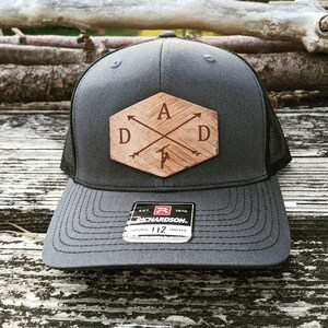 K&ouml;nnte beinhalten: Eine graue und schwarze Trucker-Kappe mit einem Holz-Patch, auf dem "DAD" mit Pfeilen steht, die auf die Buchstaben zeigen. Der Patch hat eine sechseckige Form mit einer braunen Holzmaserung. Die Kappe hat ein schwarzes Netzr&uuml;cken und ein schwarzes Schild. Die Kappe hat ein schwarzes und rotes Etikett mit der Aufschrift "Richardson" und "Original 112 Trucker".