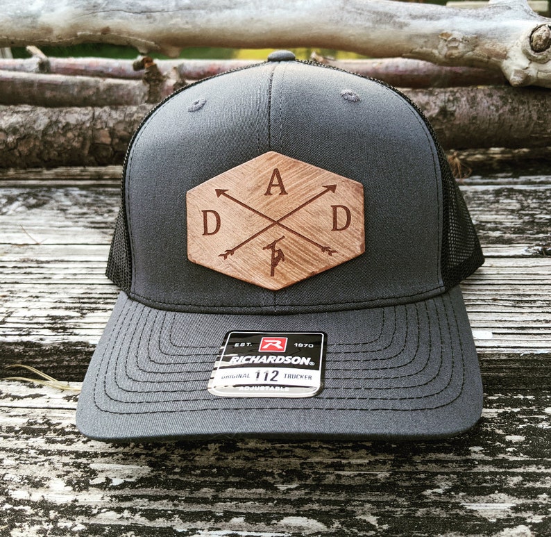 K&ouml;nnte beinhalten: Eine graue und schwarze Trucker-Kappe mit einem Holz-Patch, auf dem "DAD" mit Pfeilen steht, die auf die Buchstaben zeigen. Der Patch hat eine sechseckige Form mit einer braunen Holzmaserung. Die Kappe hat ein schwarzes Netzr&uuml;cken und ein schwarzes Schild. Die Kappe hat ein schwarzes und rotes Etikett mit der Aufschrift "Richardson" und "Original 112 Trucker".