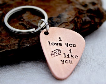 May include: A copper-colored guitar pick-shaped keychain with the words "i love you AND i like you" stamped on it. The keychain has a silver ring and is resting on a textured, dark brown surface.