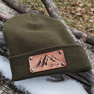 May include: A dark green beanie with a brown leather patch featuring a mountain range design and the text "Rift Canyon".