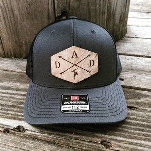 K&ouml;nnte beinhalten: Eine schwarze und graue Trucker-Kappe mit einem braunen Holz-Patch, auf dem "DAD" mit Pfeilen steht, die auf die Buchstaben zeigen. Der Patch hat die Form eines Sechsecks. Die Kappe hat ein schwarzes Netzr&uuml;cken und einen schwarzen verstellbaren Riemen mit einem roten "R"-Logo und dem Text "EST. 1970 RICHARDSON. ORIGINAL 112 TRUCKER ADJUSTABLE".