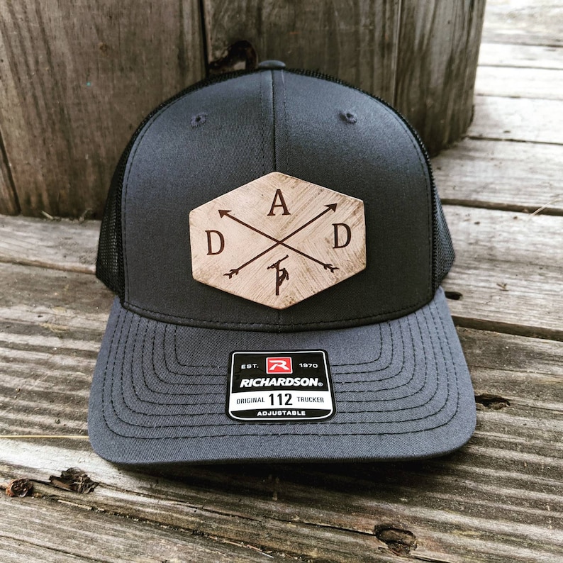 K&ouml;nnte beinhalten: Eine schwarze und graue Trucker-Kappe mit einem braunen Holz-Patch, auf dem "DAD" mit Pfeilen steht, die auf die Buchstaben zeigen. Der Patch hat die Form eines Sechsecks. Die Kappe hat ein schwarzes Netzr&uuml;cken und einen schwarzen verstellbaren Riemen mit einem roten "R"-Logo und dem Text "EST. 1970 RICHARDSON. ORIGINAL 112 TRUCKER ADJUSTABLE".