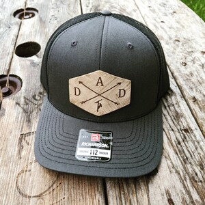 K&ouml;nnte beinhalten: Ein schwarzer und grauer Trucker-Hut mit einem braunen Holz-Patch, auf dem "DAD" mit Pfeilen steht, die auf die Buchstaben zeigen. Der Hut ist von Richardson und ist verstellbar.