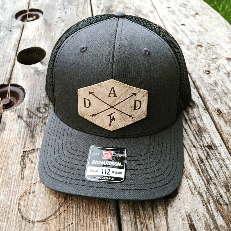 K&ouml;nnte beinhalten: Ein schwarzer und grauer Trucker-Hut mit einem braunen Holz-Patch, auf dem "DAD" mit Pfeilen steht, die auf die Buchstaben zeigen. Der Hut ist von Richardson und ist verstellbar.