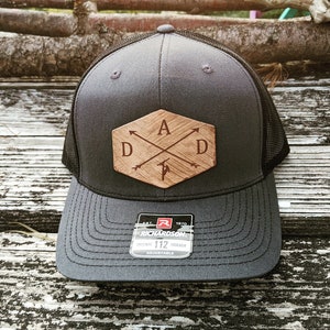 K&ouml;nnte beinhalten: Eine graue und schwarze Trucker-Kappe mit einem Holz-Patch, auf dem "DAD" mit Pfeilen und einer kleinen Axt-Grafik steht. Die Kappe ist verstellbar und tr&auml;gt das Logo der Marke Richardson.