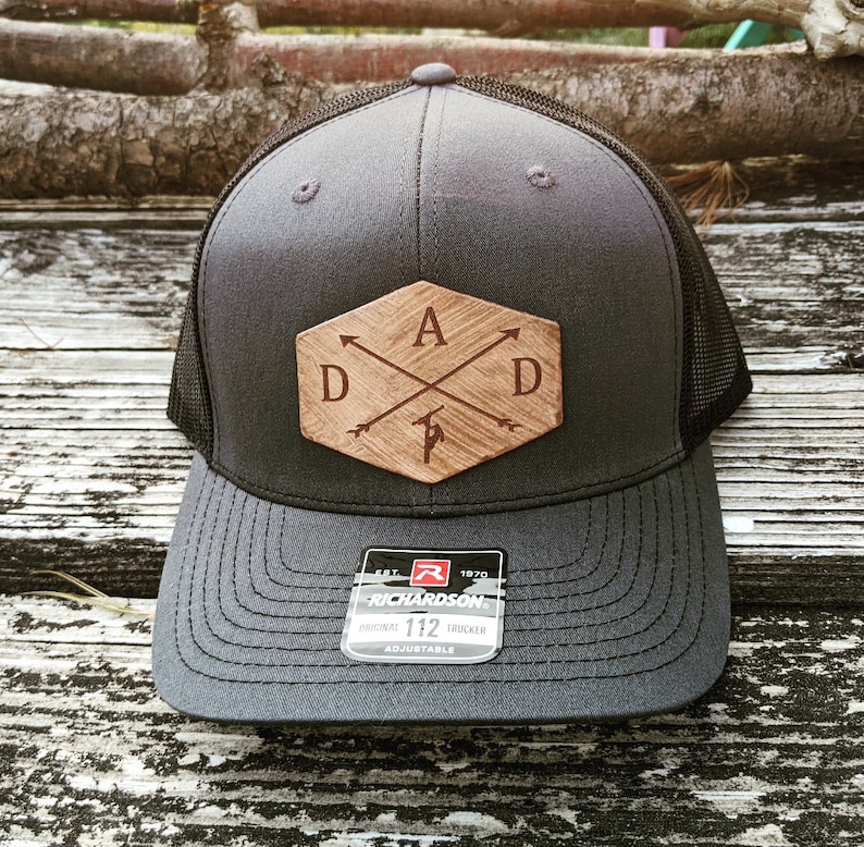K&ouml;nnte beinhalten: Eine graue und schwarze Trucker-Kappe mit einem Holz-Patch, auf dem "DAD" mit Pfeilen und einer kleinen Axt-Grafik steht. Die Kappe ist verstellbar und tr&auml;gt das Logo der Marke Richardson.