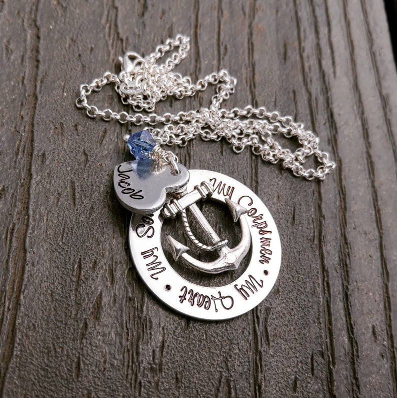May include: Silver necklace with a circular pendant featuring an anchor design. The pendant is inscribed with the words "My Sailor, My Heart, My Corpsman". A heart-shaped charm with the name "Jacob" and a blue crystal are also included.