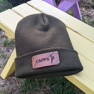 May include: Olive green beanie hat with a brown leather patch. The patch is riveted to the hat and features the word "CAPPS" and a graphic. The hat is made of a knitted material.
