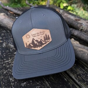 May include: A gray and black trucker hat with a wooden patch that says "Leave the Road Take the Trail". The patch features a mountain scene with trees and a sun.
