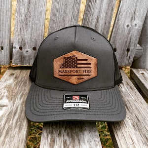 May include: A black and gray trucker hat with a wood patch featuring an American flag and the text "MASSPORT FIRE". The hat is adjustable and has a black mesh back.