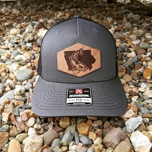 May include: A gray and black trucker hat with a brown leather patch featuring a catfish and the outline of a state. The patch is sewn onto the front of the hat. The hat has an adjustable strap and a black mesh back. The hat has the text "EST. 1970 RICHARDSON. ORIGINAL TRUCKER 112 ADJUSTABLE" on the back.