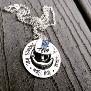 May include: A silver necklace with a pendant featuring an anchor design. The pendant has the words "My Son, My Heart, Seabee" around the edge. A heart-shaped charm with the name "Tyler" is attached, along with a blue crystal.