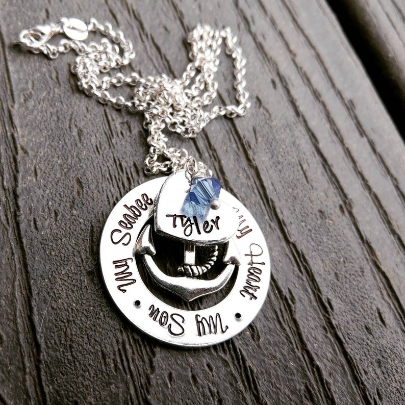 May include: A silver necklace with a pendant featuring an anchor design. The pendant has the words "My Son, My Heart, Seabee" around the edge. A heart-shaped charm with the name "Tyler" is attached, along with a blue crystal.